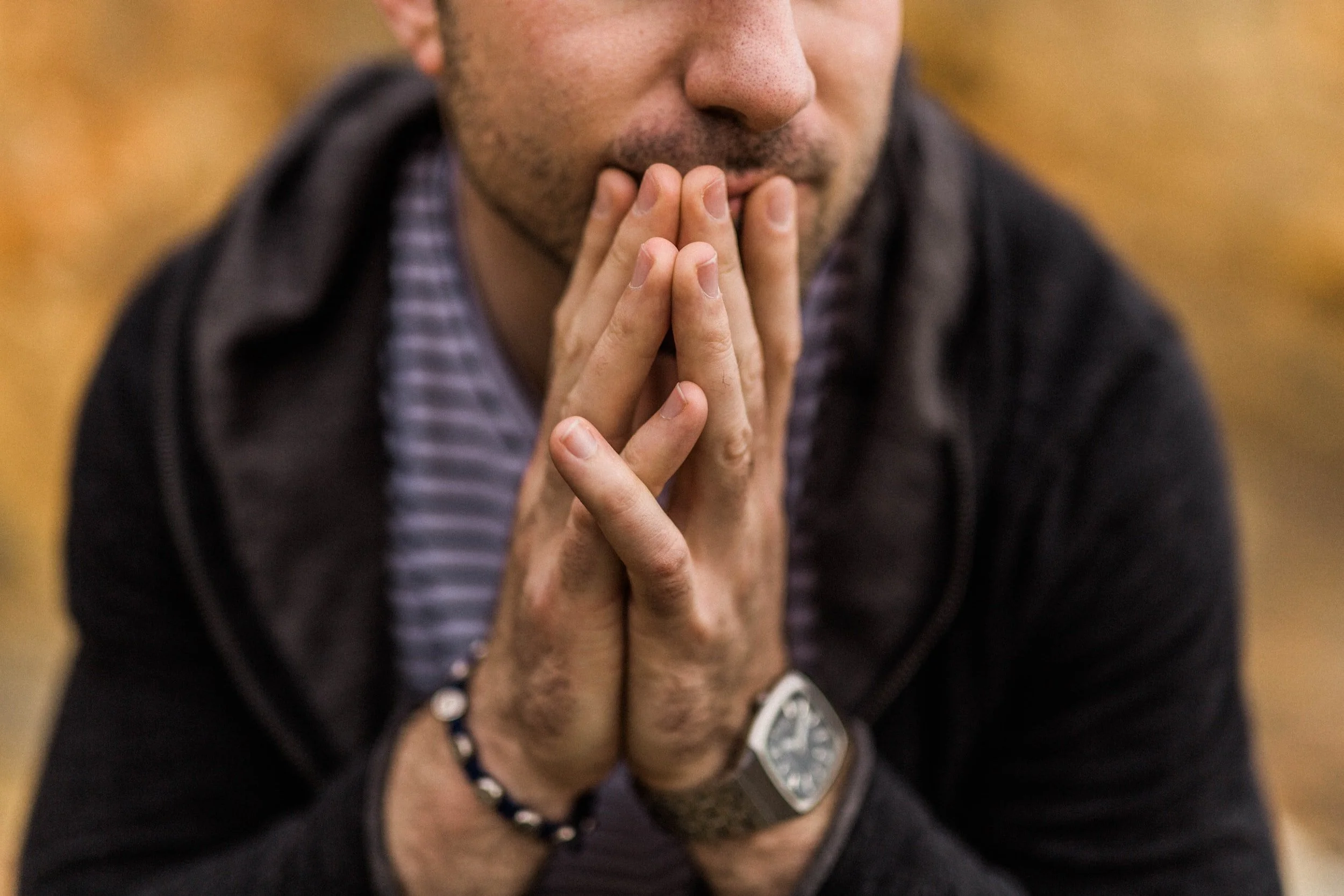 Man with hands clasped, deep in thought, wearing a watch and bracelet.