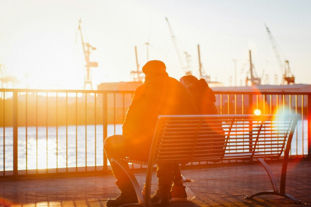 Couple sitting on a bench at sunset, overlooking a harbor with cranes.