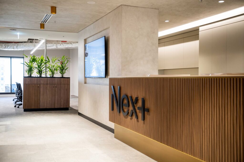 Modern office reception desk with "Nex+" logo, plants, and screen.