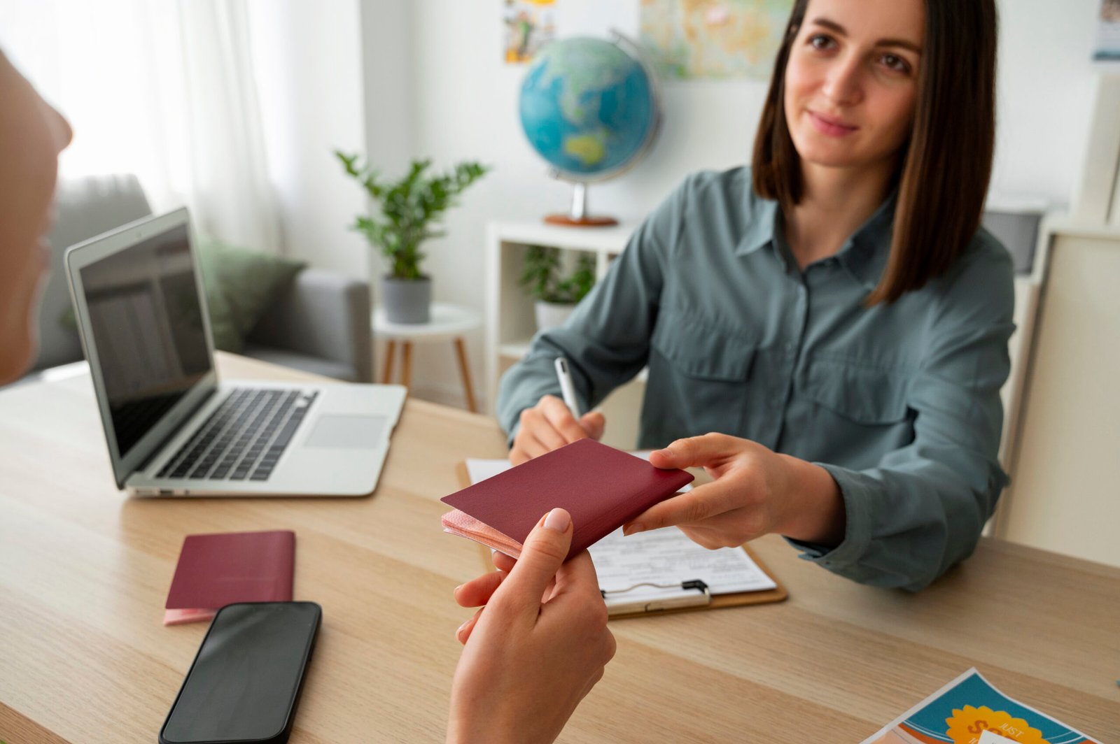 Woman handing a passport to someone across a desk with a laptop and potted plant