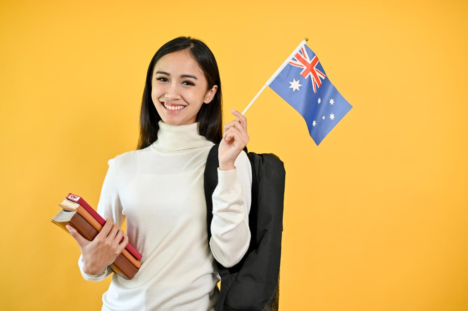 Smiling student with Australian flag, books, and backpack against a yellow background.