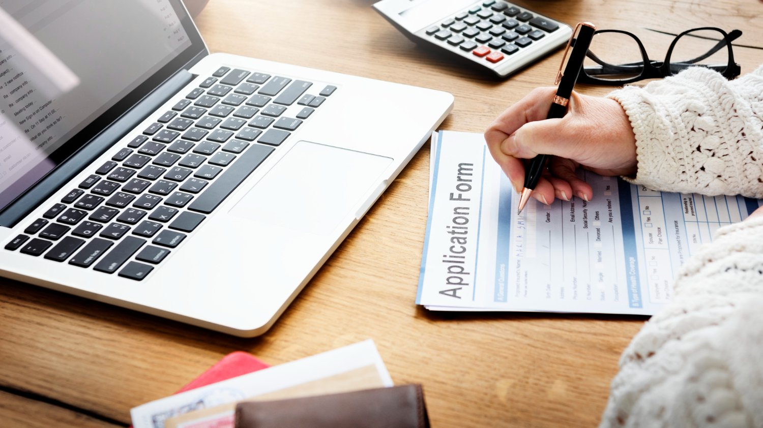 Filling out an application form with a laptop, calculator, and glasses on a wooden desk. "Application Form" text visible.